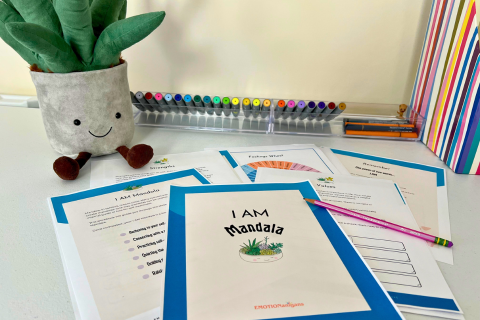A bright workspace features a smiling plush aloe vera plant, lined-up colorful pens, and a stack of worksheets titled "I AM Mandala" on a white desk. A pink pencil lies on top, ready for use.