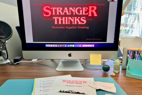 A desktop computer screen displays a presentation titled "Stranger Thinks: Neutralize Negative Thinking" in a similar style to the Stranger Things logo. On the desk are documents, a microphone, and a 'Stranger Thinks' therapy guide booklet and tool cards.