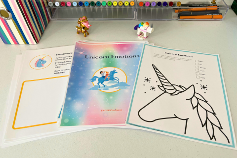 A desk with unicorn-themed coloring and activity sheets. The main booklet titled "Unicorn Emotions" features a colorful cover with a unicorn with 2 children on its back. The desk also has markers, pens, a small unicorn figurine, and a striped organizer.